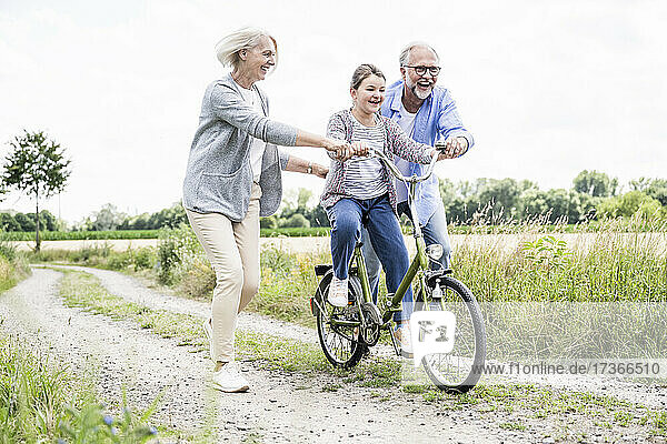 Cheerful grandparent helping granddaughter while riding bicycle on dirt road
