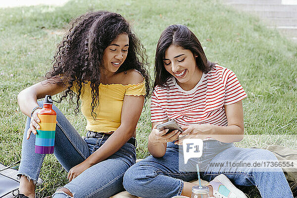 Junge Frau sieht ihre Freundin an  die ein Smartphone benutzt  während sie im Park sitzt