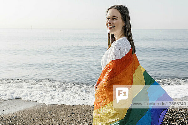 Lächelnde Frau in Regenbogenfahne eingewickelt am Strand stehend