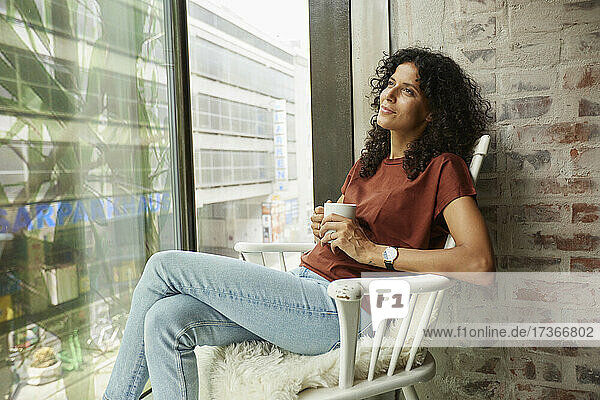 Nachdenkliche Frau mit Kaffeetasse auf einem Stuhl am Fenster sitzend