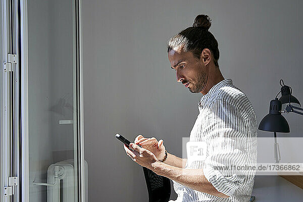 Männlicher Berufstätiger  der ein Mobiltelefon vor einem Fenster im Büro benutzt