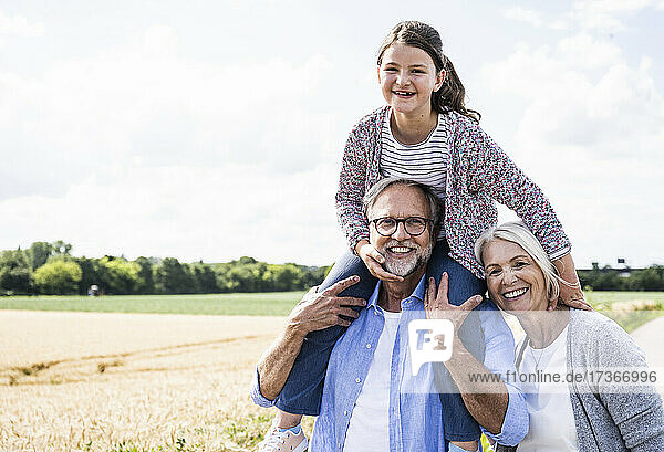 Fröhlicher Mann  der seine Enkelin auf der Schulter trägt  während die Frau mit ihm einen sonnigen Tag verbringt