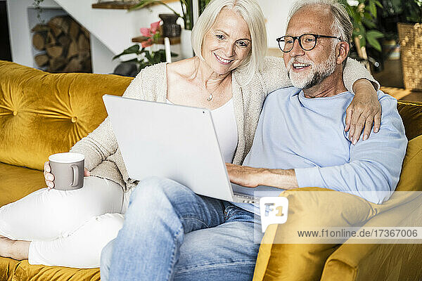 Lächelnde reife Frau,  die den Arm um einen Mann legt,  der zu Hause einen Laptop benutzt