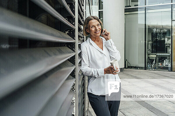 Lächelnde Geschäftsfrau  die einen Kaffee trinkt  während sie mit einem Mobiltelefon im Büropark spricht