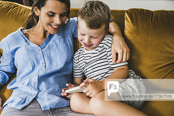 Frau mit Arm um Sohn  der auf dem Sofa zu Hause sein Smartphone benutzt