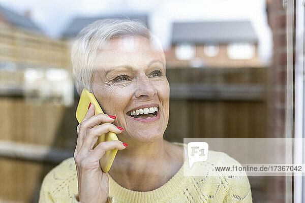 Glückliche reife Frau  die mit einem Smartphone spricht und durch ein Glasfenster schaut