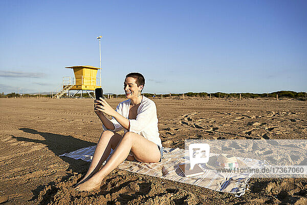 Lächelnde Frau  die ein Mobiltelefon benutzt  während sie am Strand sitzt