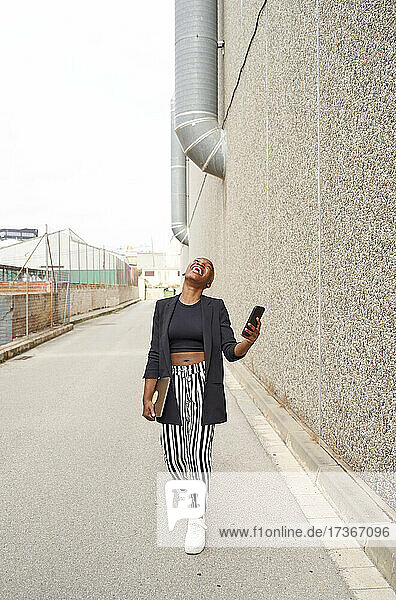 Geschäftsfrau lachend beim Gehen mit Smartphone und Laptop auf dem Fußweg