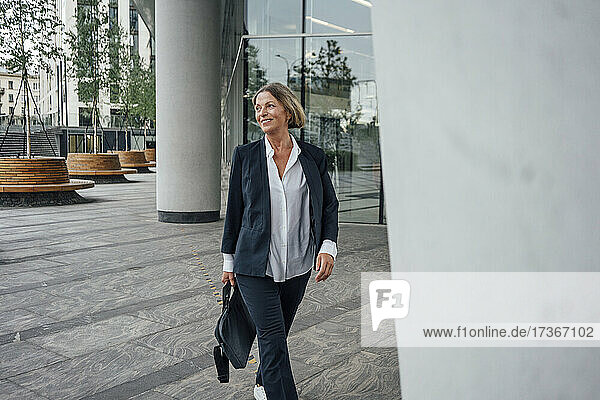 Smiling female professional with bag looking away while walking on walkway