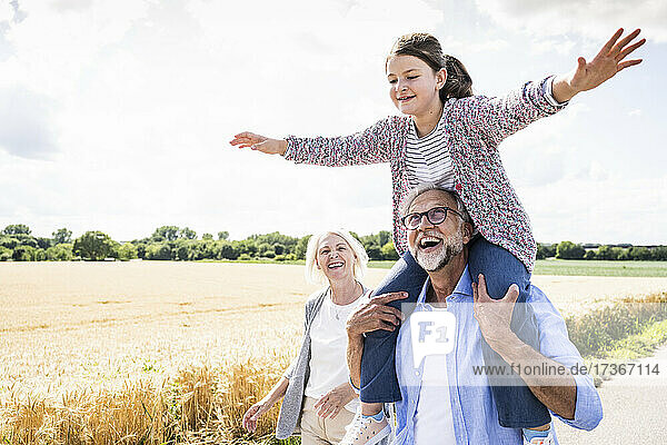 Glücklicher Mann  der seine Enkelin auf der Schulter trägt  während er an einem sonnigen Tag auf der Straße spazieren geht