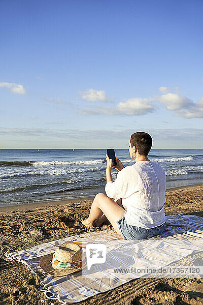 Frau fotografiert am Strand sitzend das Meer mit dem Handy