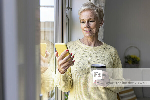 Reife Frau  die ein Smartphone benutzt und eine Tasse zu Hause hält