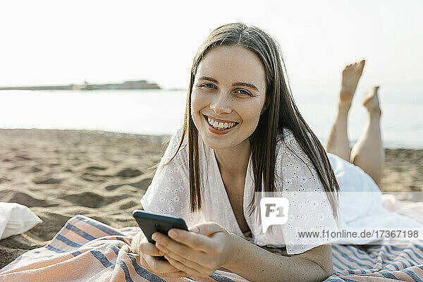 Smiling woman holding mobile phone while relaxing at beach