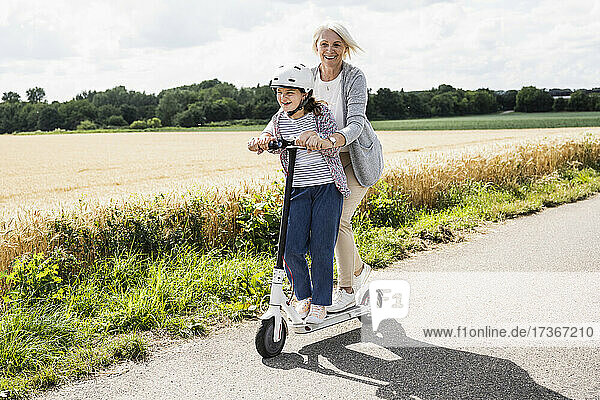 Fröhliches Mädchen fährt Roller mit Frau auf der Straße an einem sonnigen Tag