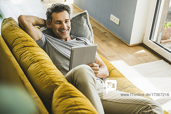 Lächelnder Mann  der ein digitales Tablet benutzt  während er zu Hause auf dem Sofa liegt