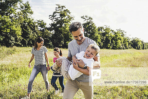 Lächelnder Vater trägt seinen Sohn beim Spaziergang mit der Familie auf einer Wiese