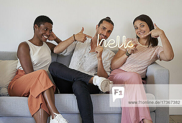Glückliche männliche und weibliche Mitarbeiter  die auf dem Sofa sitzend auf das Hello-Zeichen mit Leuchtschrift zeigen