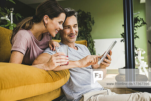Frau hält ein Saftglas  während ein Mann ein digitales Tablet zu Hause benutzt
