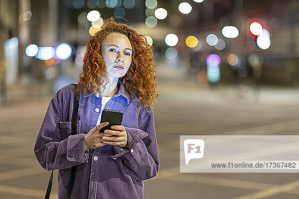 Rothaarige Frau mit Mobiltelefon  die nachts auf einer Kreuzung steht und wegschaut