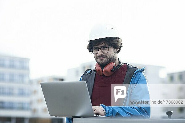 Junger Ingenieur mit Helm und Hörschutz checkt mit Laptop Arbeitstelle außen  Freiburg  Baden-Württemberg  Deutschland  Europa