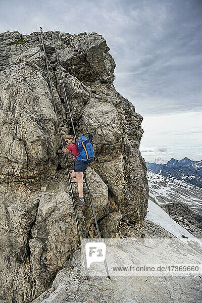 Wanderin besteigt Metalleiter am Fels  Bergsteiger auf Gratweg mit Schneeresten  dramatischer Wolkenhimmel  Heilbronner Weg  Allgäuer Alpen  Allgäu  Bayern  Deutschland  Europa