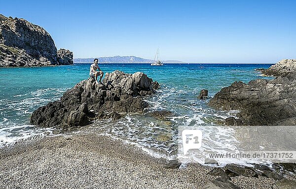 Junger Mann sitzt auf einem Felsen  Strand mit Vulkanfelsen  türkisblaues Meer  hinten Segel-Katamaran  Gyali  Dodekanes  Griechenland  Europa