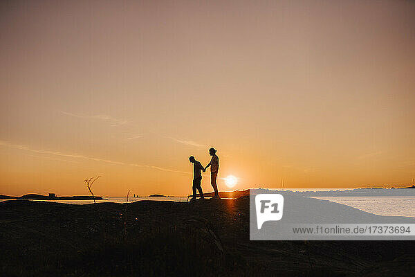 Männliche Freunde halten sich beim Spaziergang am Seeufer während des Sonnenuntergangs an den Händen
