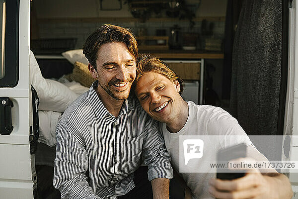 Smiling male friends taking selfie on mobile phone at doorway of camping van