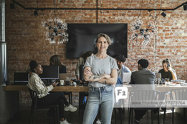 Female entrepreneur standing with arms crossed while colleagues working in creative office