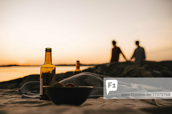 Bierflasche und Schüssel auf Picknickdecke  während schwules Paar im Hintergrund am Seeufer bei Sonnenuntergang