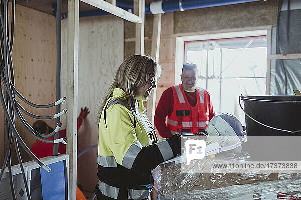 Lächelnde Bauunternehmerin  die einen Grundriss analysiert  während sie sich mit einem männlichen Kollegen auf der Baustelle unterhält