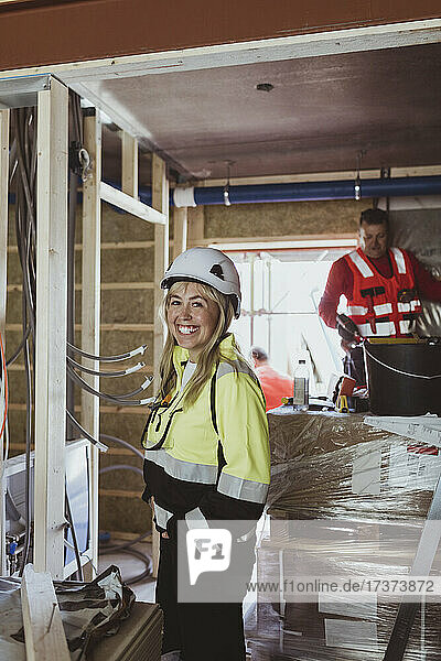 Happy female building contractor standing with hand in pocket while male construction worker working at site