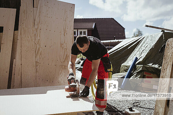 Männlicher Zimmermann  der an einem sonnigen Tag auf einer Baustelle ein Holzbrett mit einer elektrischen Säge schneidet