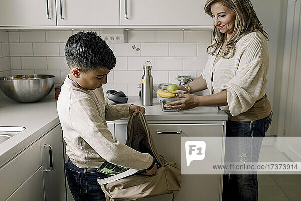 Lächelnde Mutter sieht ihren Sohn an  der in der Küche ein Buch in die Tasche steckt