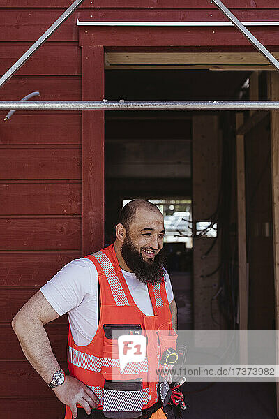 Lächelnder bärtiger männlicher Bauarbeiter  der mit der Hand auf der Hüfte vor der Tür steht