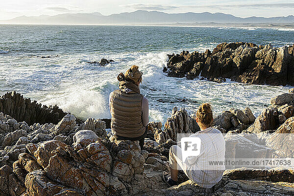 Woman and teenage girl sitting on rocky shore  looking out to sea.