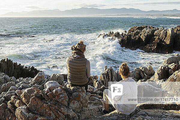 Woman and teenage girl sitting on rocky shore  looking out to sea.
