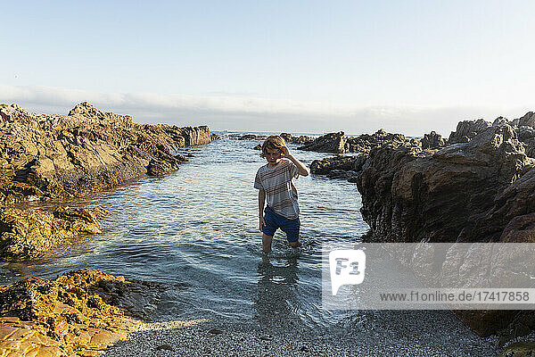 A young boy in shallow sea water among jagged rocks on the beach at De Kelders.