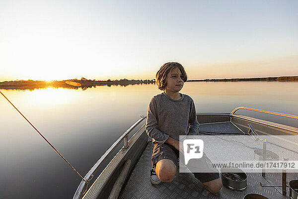 Ein Junge fischt von einem Boot aus in den flachen  ruhigen Gewässern des Okavango-Deltas bei Sonnenuntergang