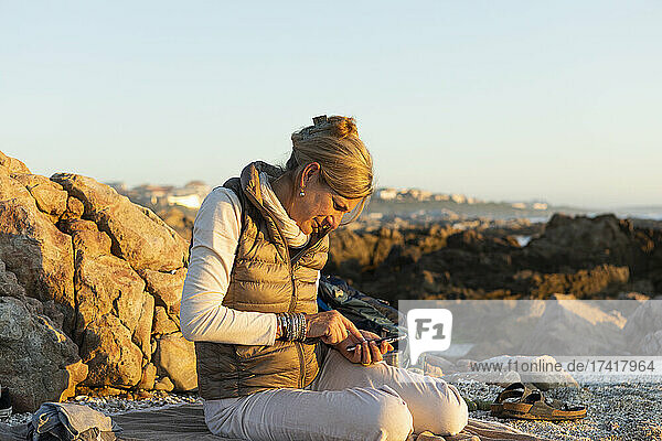 Eine erwachsene Frau sitzt bei Sonnenuntergang am Strand von De Kelders und benutzt ihr Smartphone.
