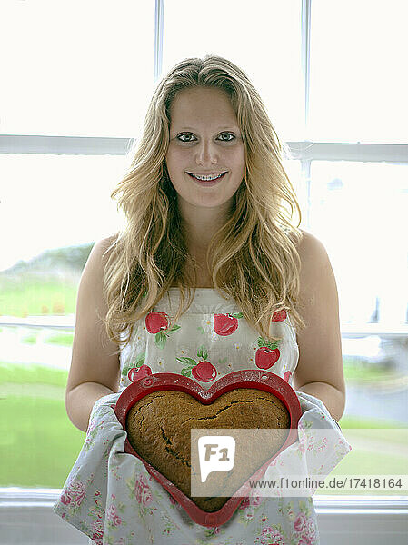 Smiling girl showing heart shaped cake while standing at home