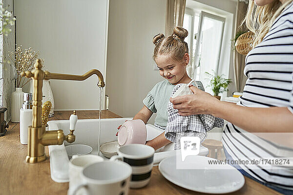 Smiling daughter washing crockery in sink with mother at home
