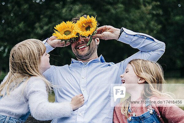 Fröhlicher Vater hält Sonnenblumen in der Hand  während er mit seiner Tochter im Park spielt