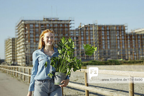 Lächelnde rothaarige Frau mit Pflanze geht an sonnigen Tagen spazieren