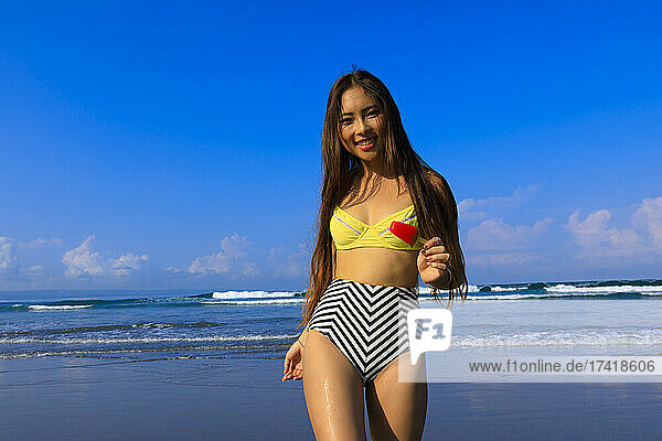 Smiling young woman wearing bikini holding ice cream at beach during sunny day