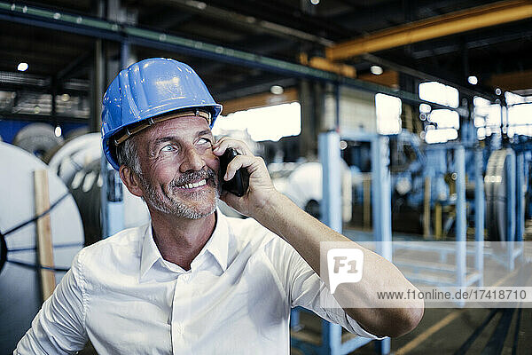 Smiling male engineer with hardhat talking on mobile phone at industry