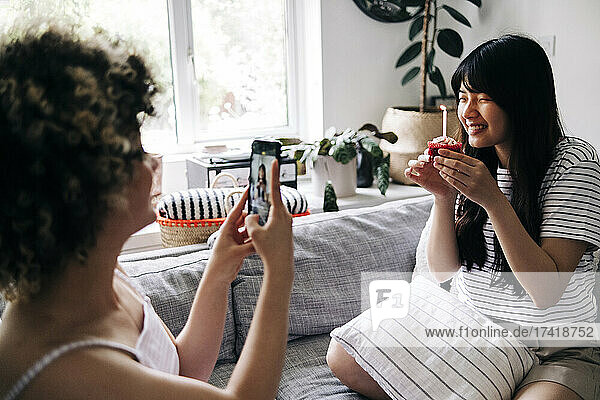 Woman taking photo of friend holding cupcake through mobile phone at home
