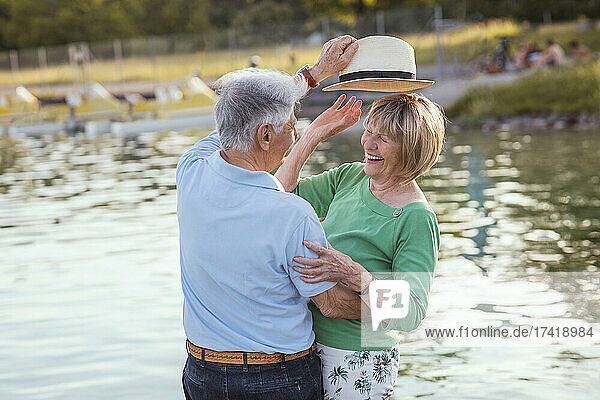 Senior man putting hat on cheerful woman by lake
