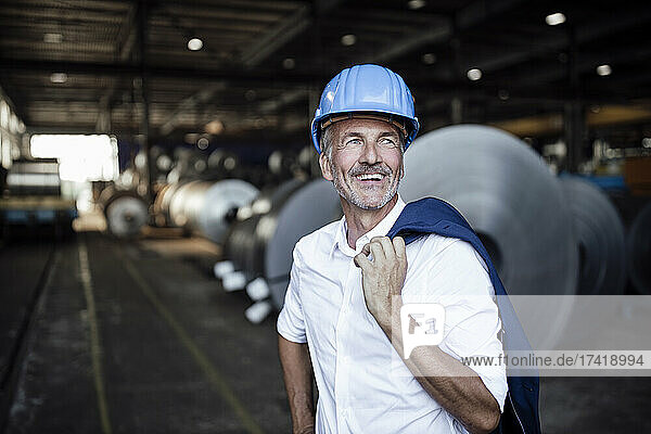 Cheerful male engineer with hardhat holding blazer at warehouse