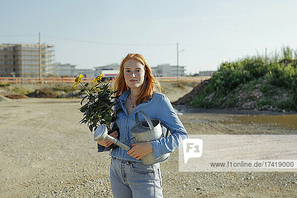 Confident woman holding potted plant and watering can at construction site